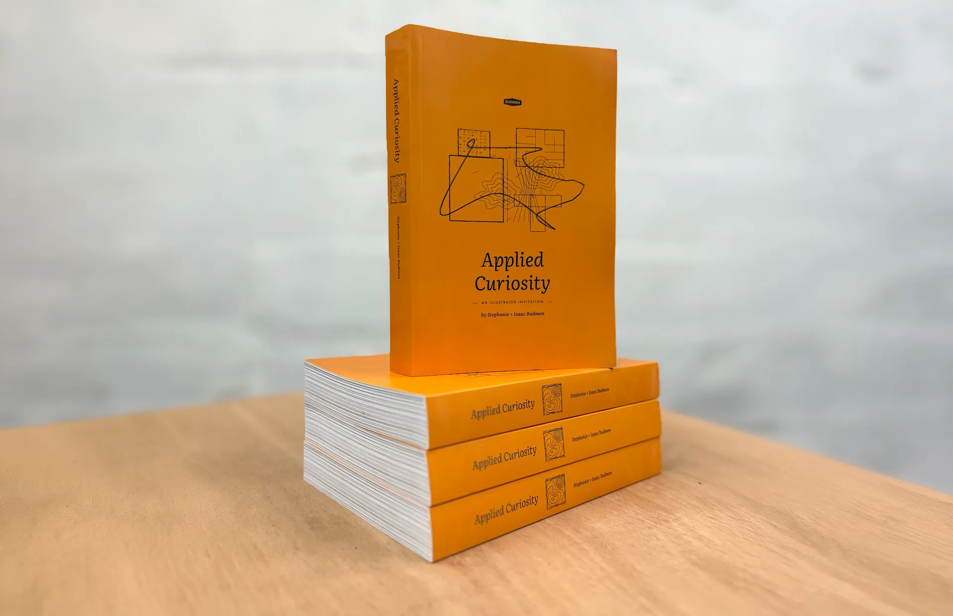 Four printed copies of Applied Curiosity on a wood table: three stacked flat showing their thick page edges, one standing upright on top with the orange cover facing out