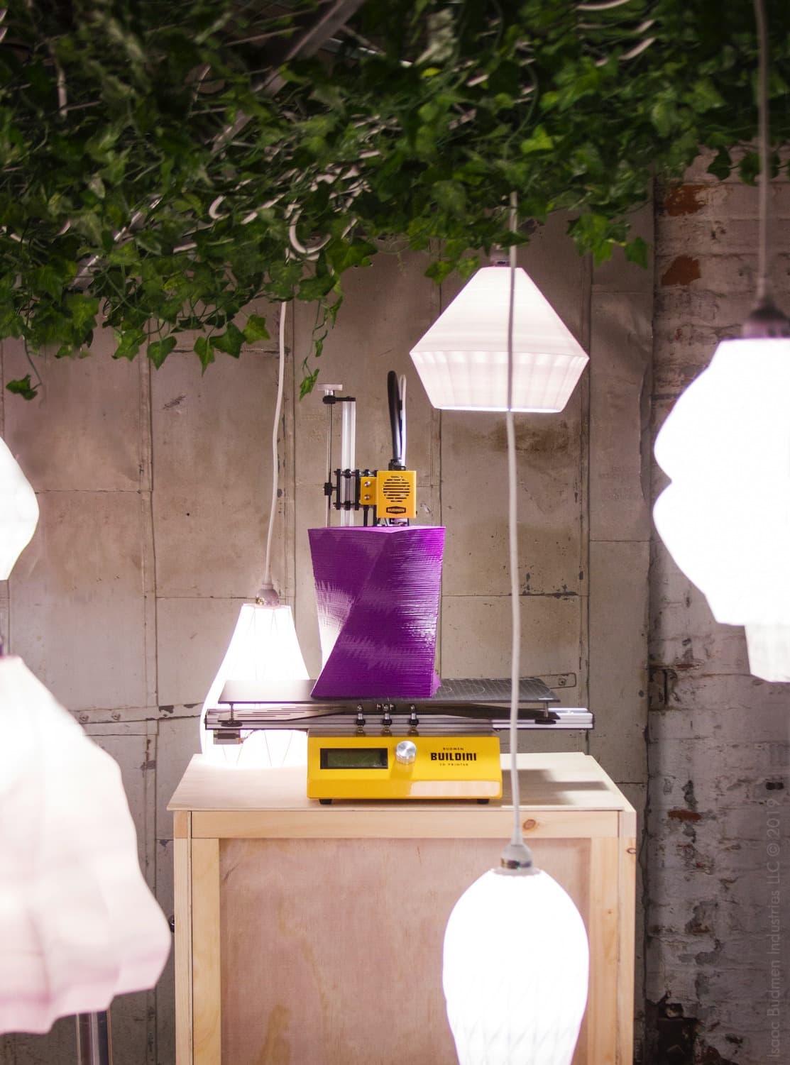 The yellow Buildini 3D printer printing a purple object surrounded by hanging lampshades