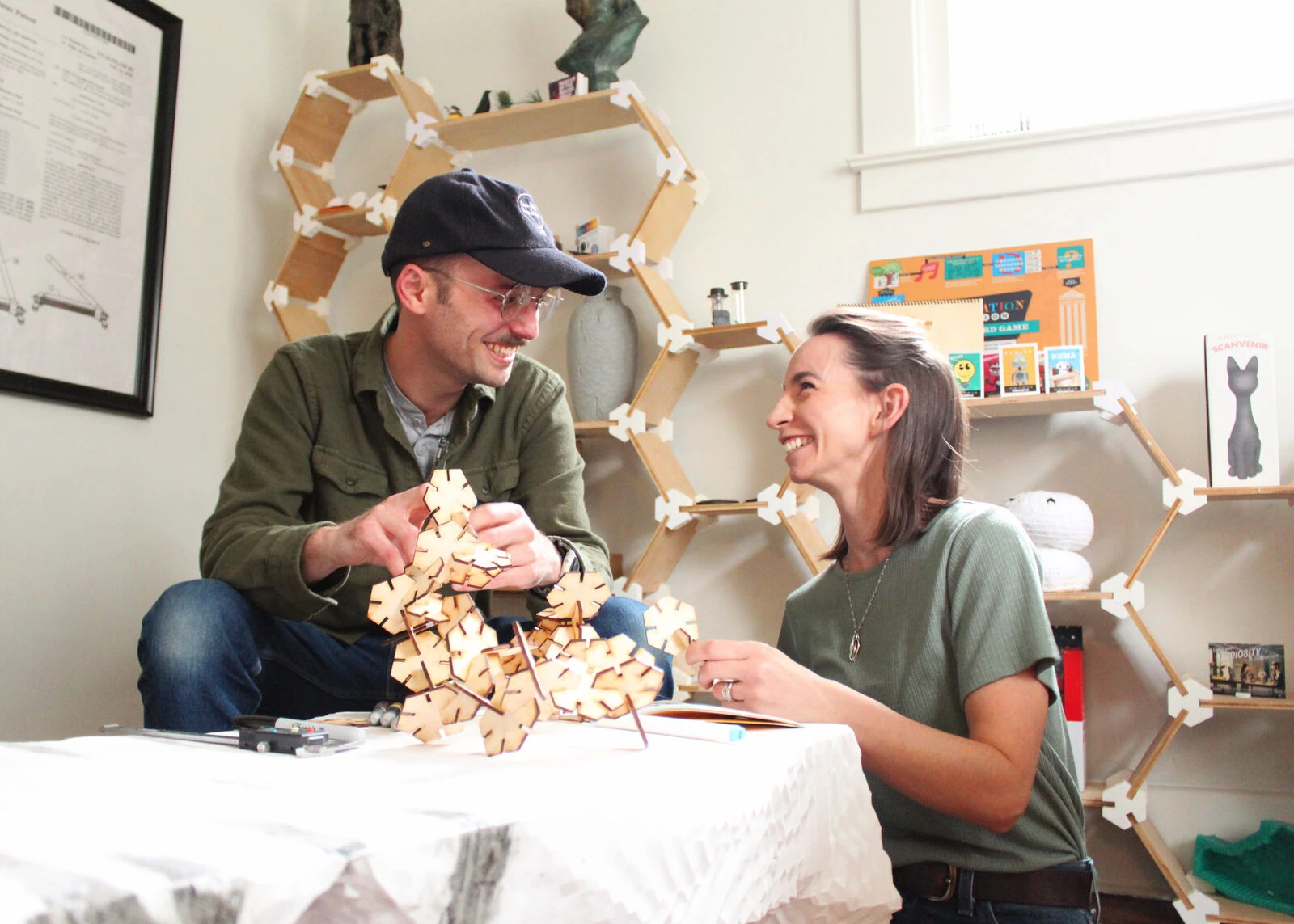Stephanie and Isaac laughing across a worktable while assembling a wooden structure in their studio