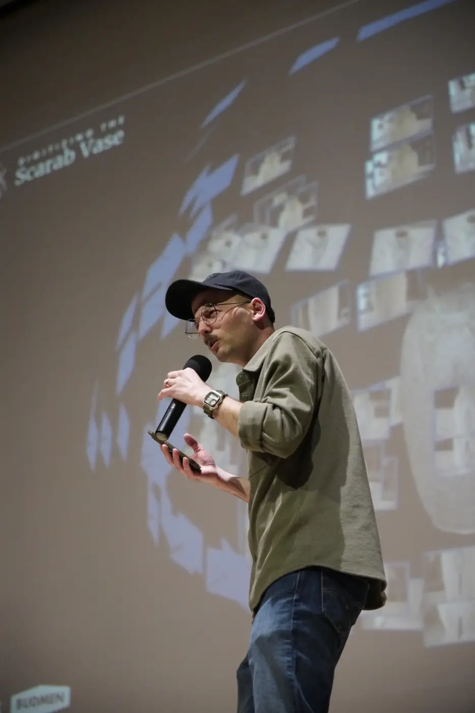 Isaac presenting on stage, microphone in hand, Scarab Vase slides projected behind him